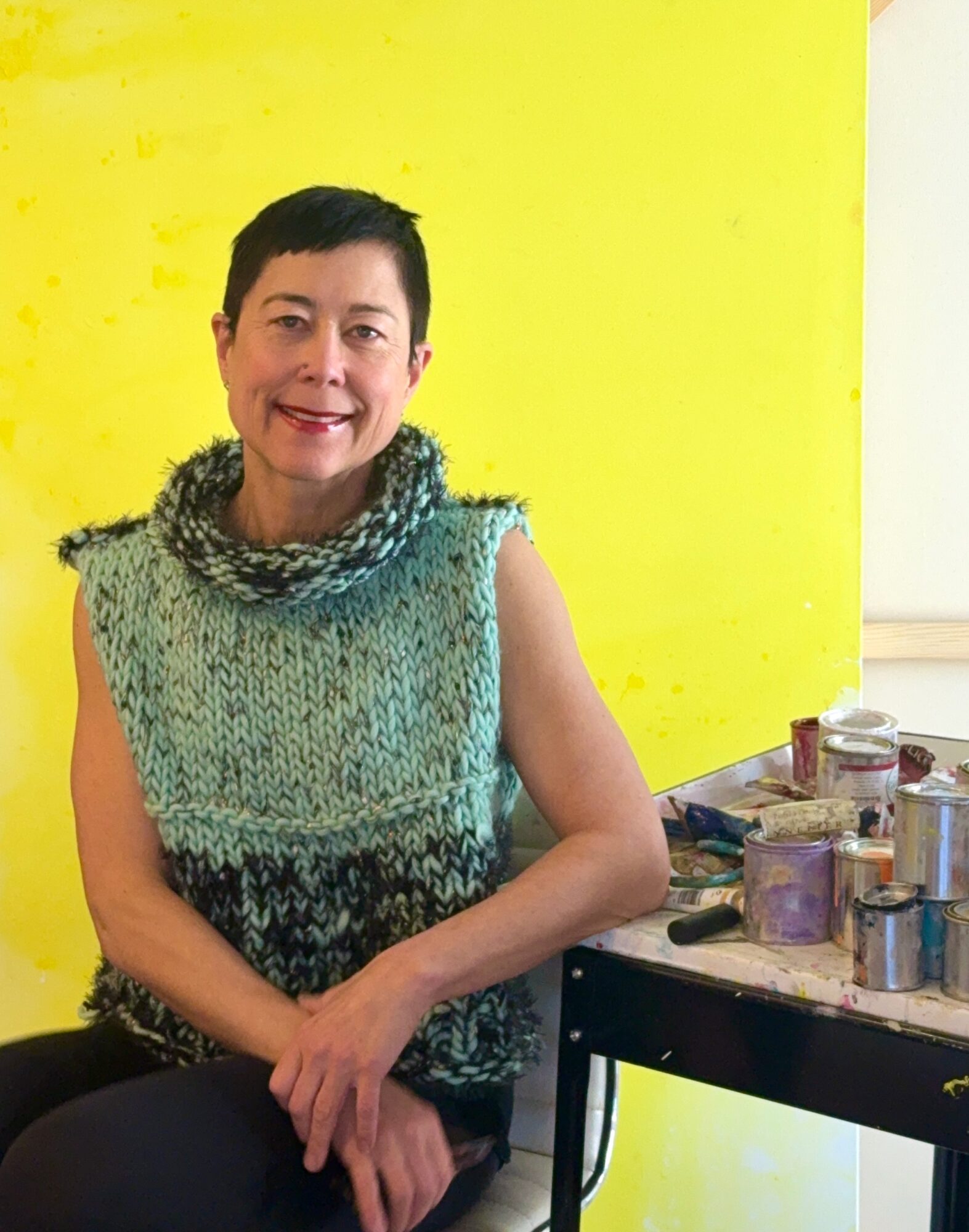 Smiling woman wearing a blue knitted scarf, seated indoors by a table.